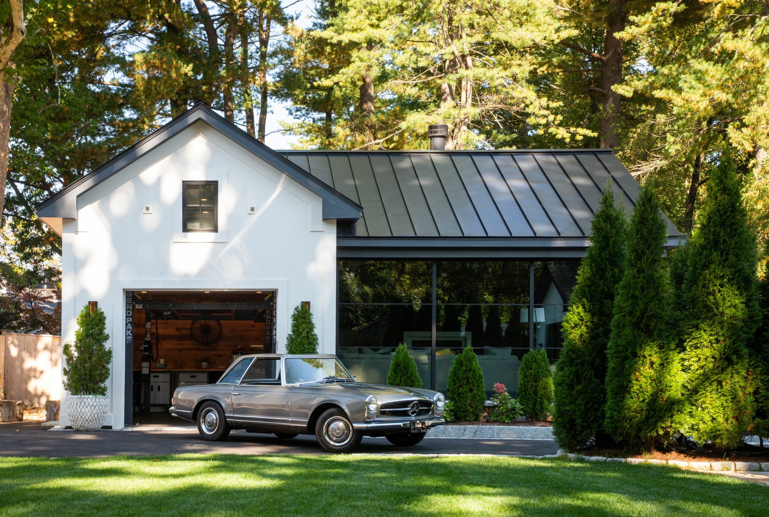 Garage_Vintage Car Modern garage addition with white exterior and black metal roof, featuring a glass-walled living space and a vintage car showcased at the entry, set within a landscaped residential setting.<br />