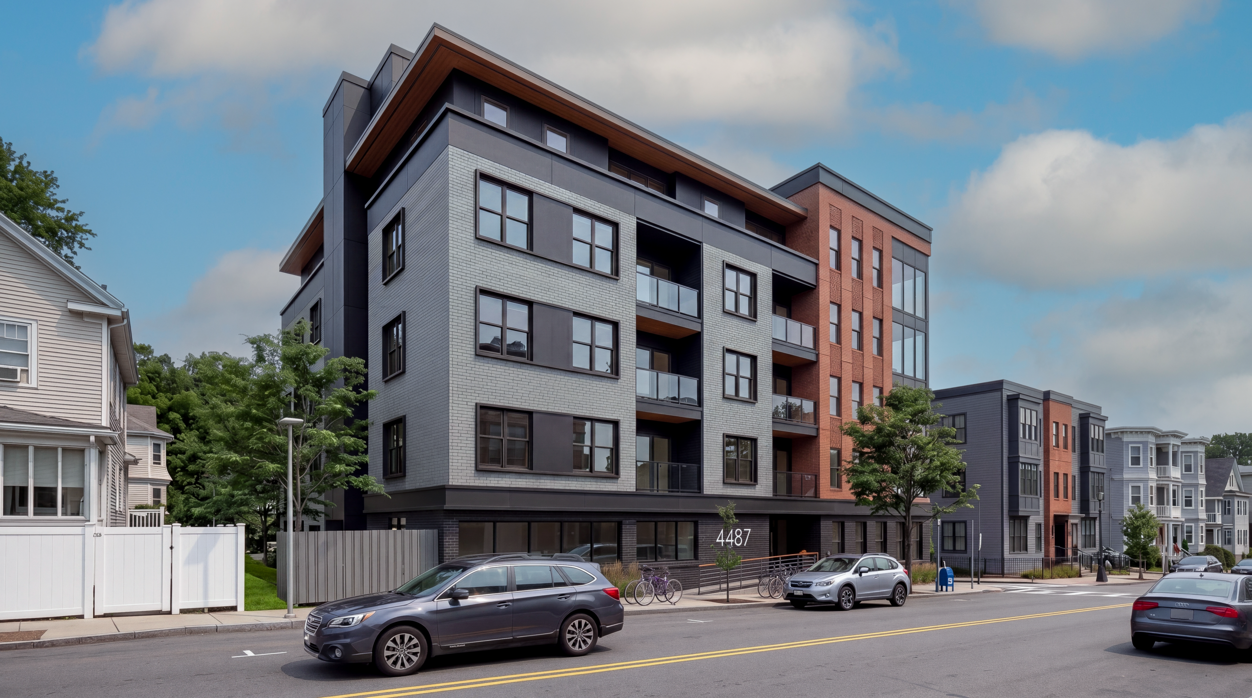 Contemporary multifamily residential building in Boston with brick and panel siding, private balconies, and street-level frontage along a tree-lined neighborhood street with parked cars