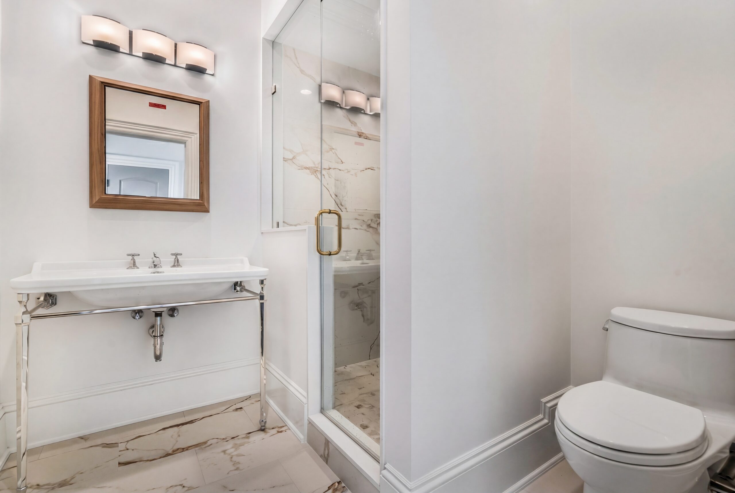 Light-filled bathroom with pedestal sink, glass-enclosed shower, marble tile flooring, and brass accents