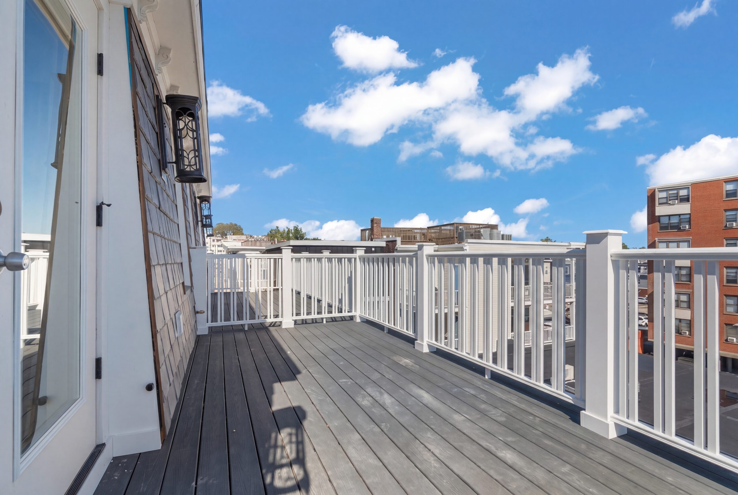 Sunlit rooftop deck with gray flooring, white railing, and surrounding neighborhood views under blue sky