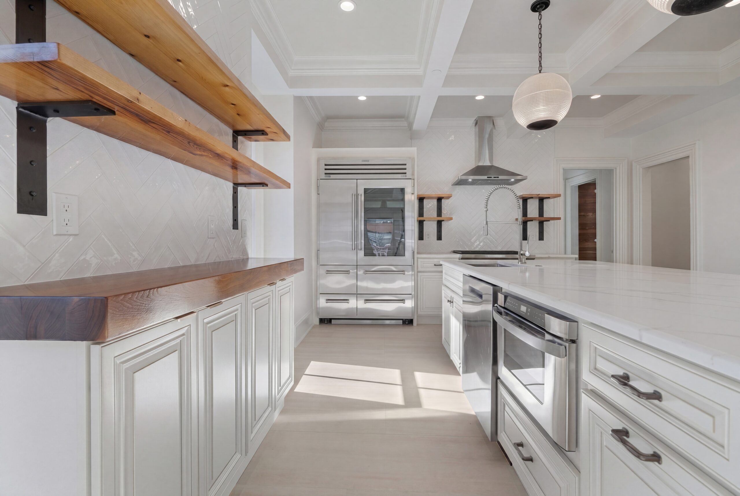 Sunlit kitchen with white cabinetry, wood open shelving, herringbone backsplash, and stainless steel appliances