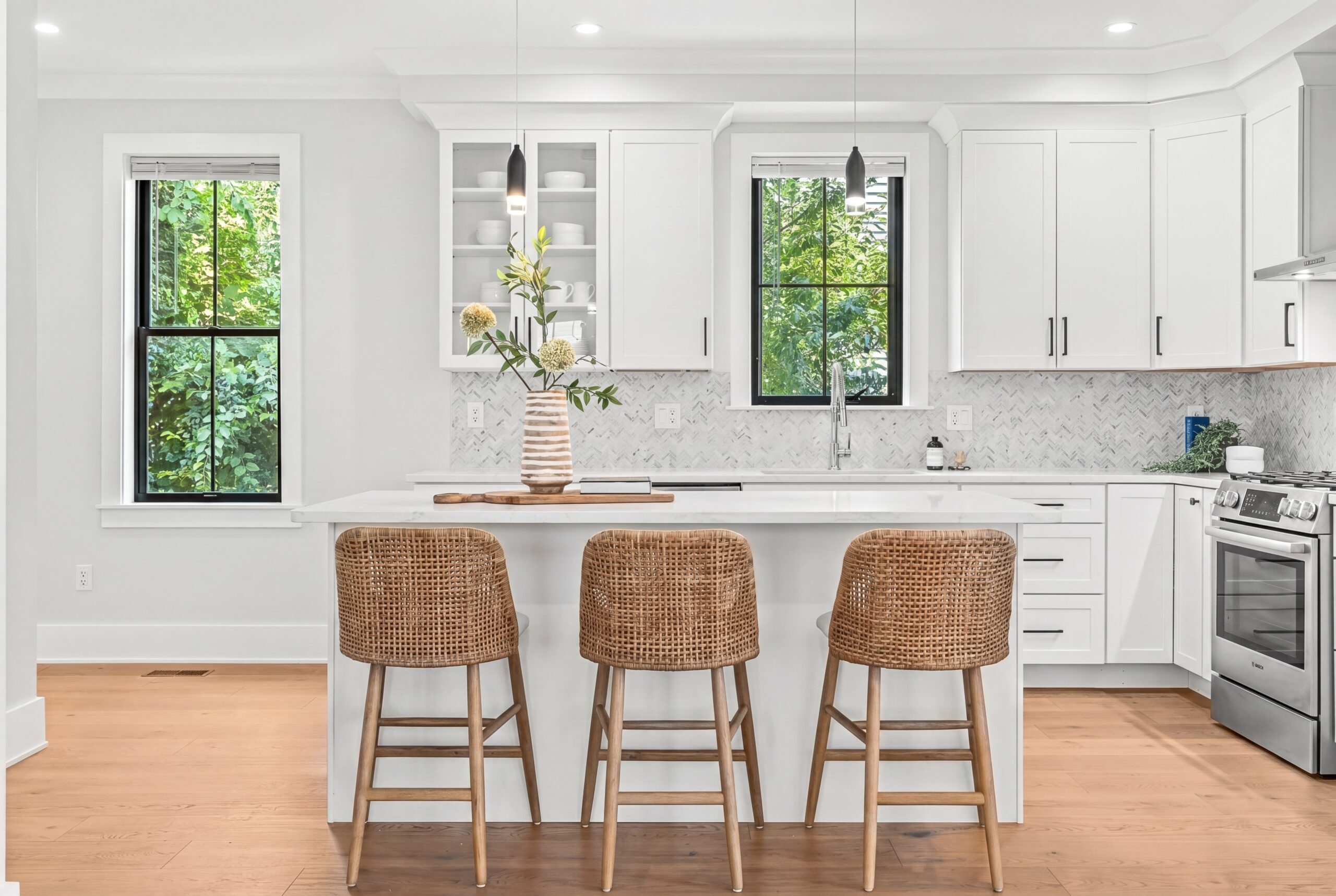 Bright kitchen with white cabinetry, waterfall island, woven bar stools, herringbone backsplash, and black-framed windows overlooking greenery