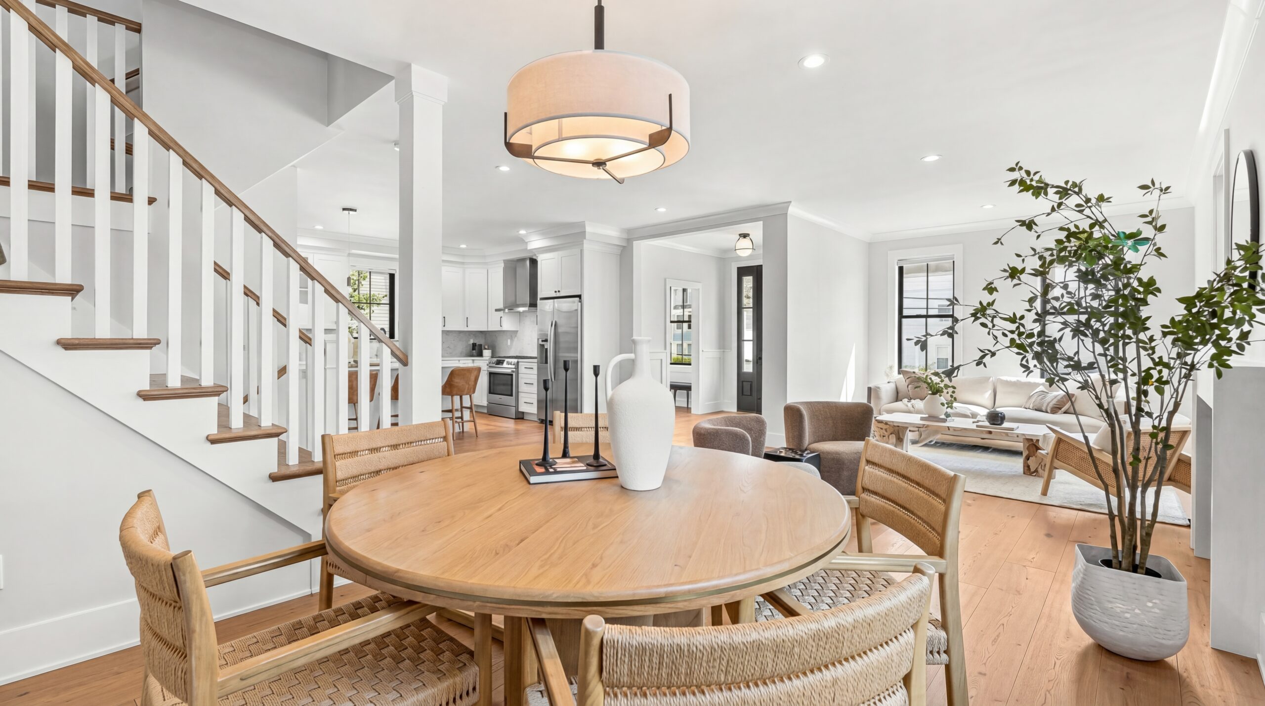 Open-concept dining and living area with round wood table, woven chairs, warm hardwood floors, staircase with white balusters, and bright kitchen beyond<br />
