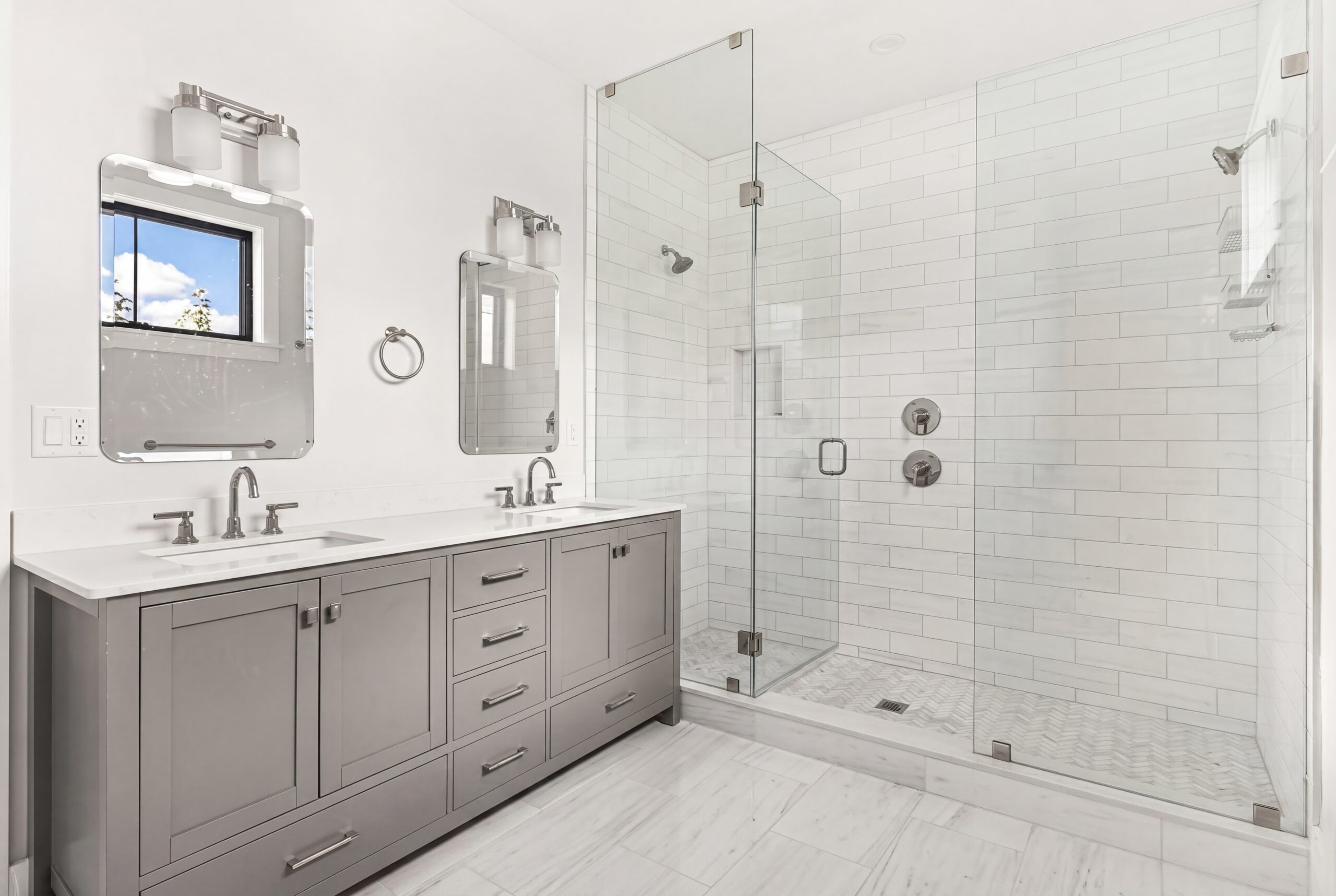 Spacious modern bathroom with double vanity, gray cabinetry, quartz countertops, and glass-enclosed walk-in shower with subway tile and marble flooring<br />
