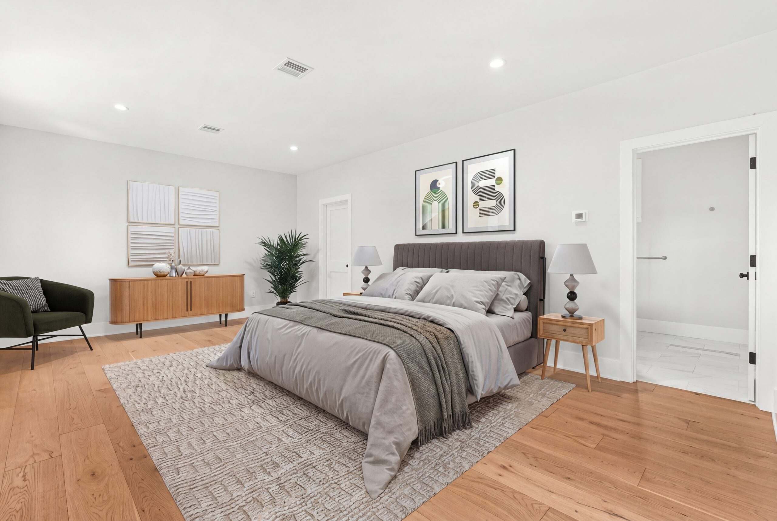 Bright and spacious primary bedroom with warm hardwood floors, upholstered bed, minimalist decor, and ensuite bath access<br />
