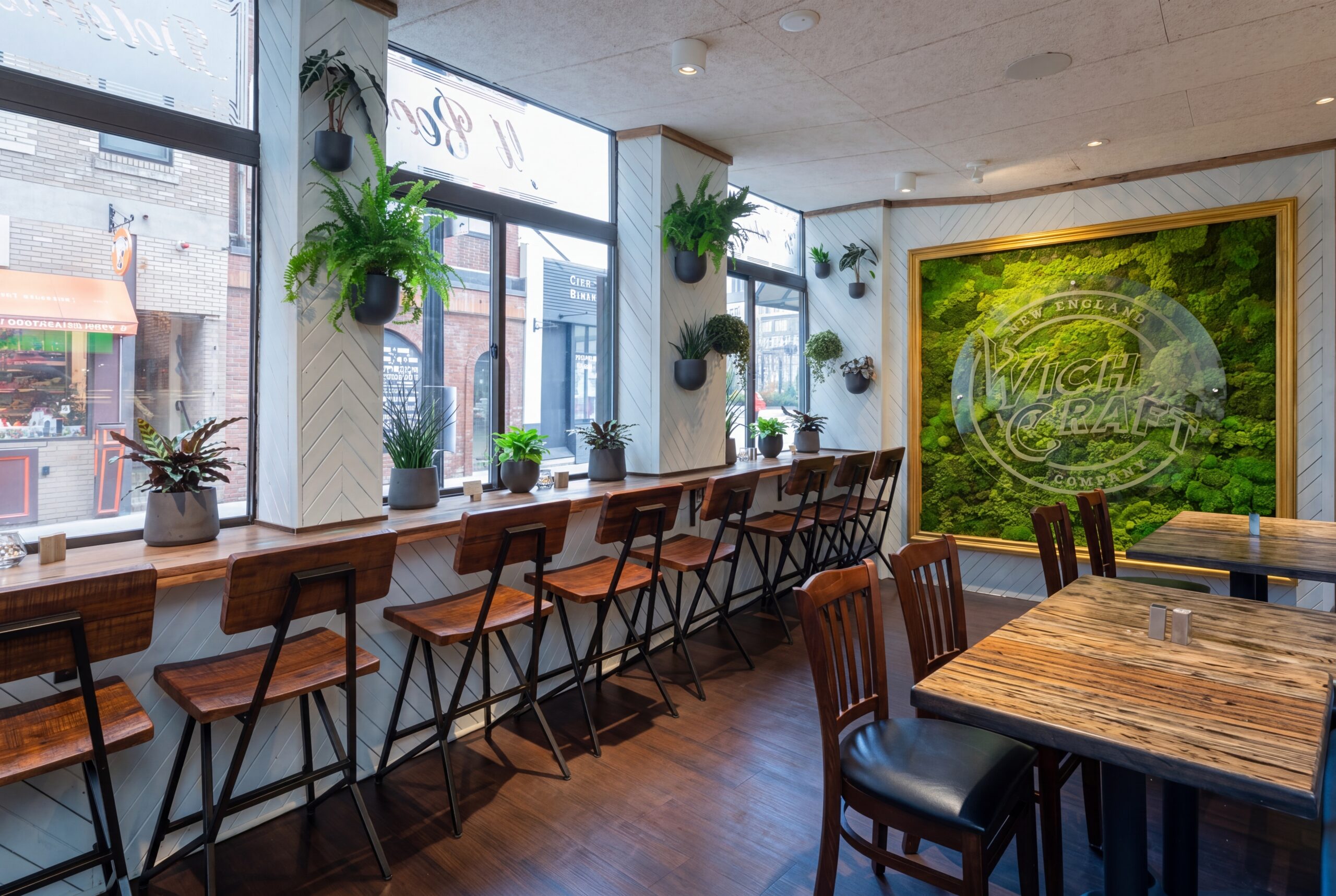 Warm café-style interior with window bar seating, wood stools and tables, hanging greenery, and a statement moss wall creating a cozy, biophilic dining atmosphere