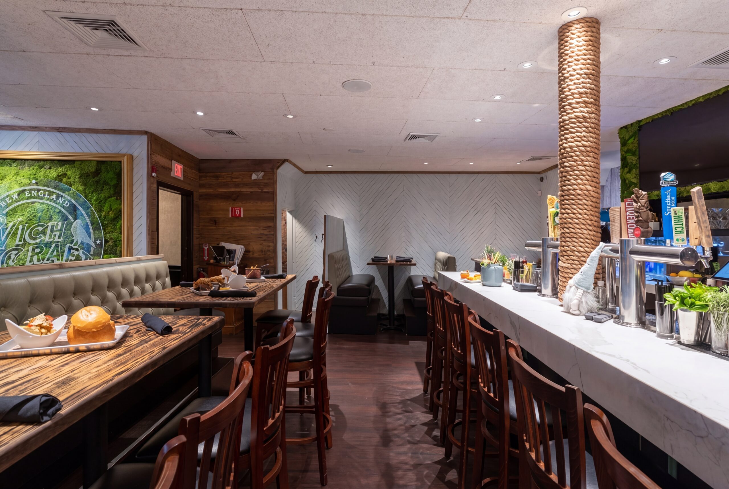 Inviting restaurant interior with bar seating, warm wood finishes, tufted booths, and a lively atmosphere anchored by craft beer taps and a signature moss wall<br />
