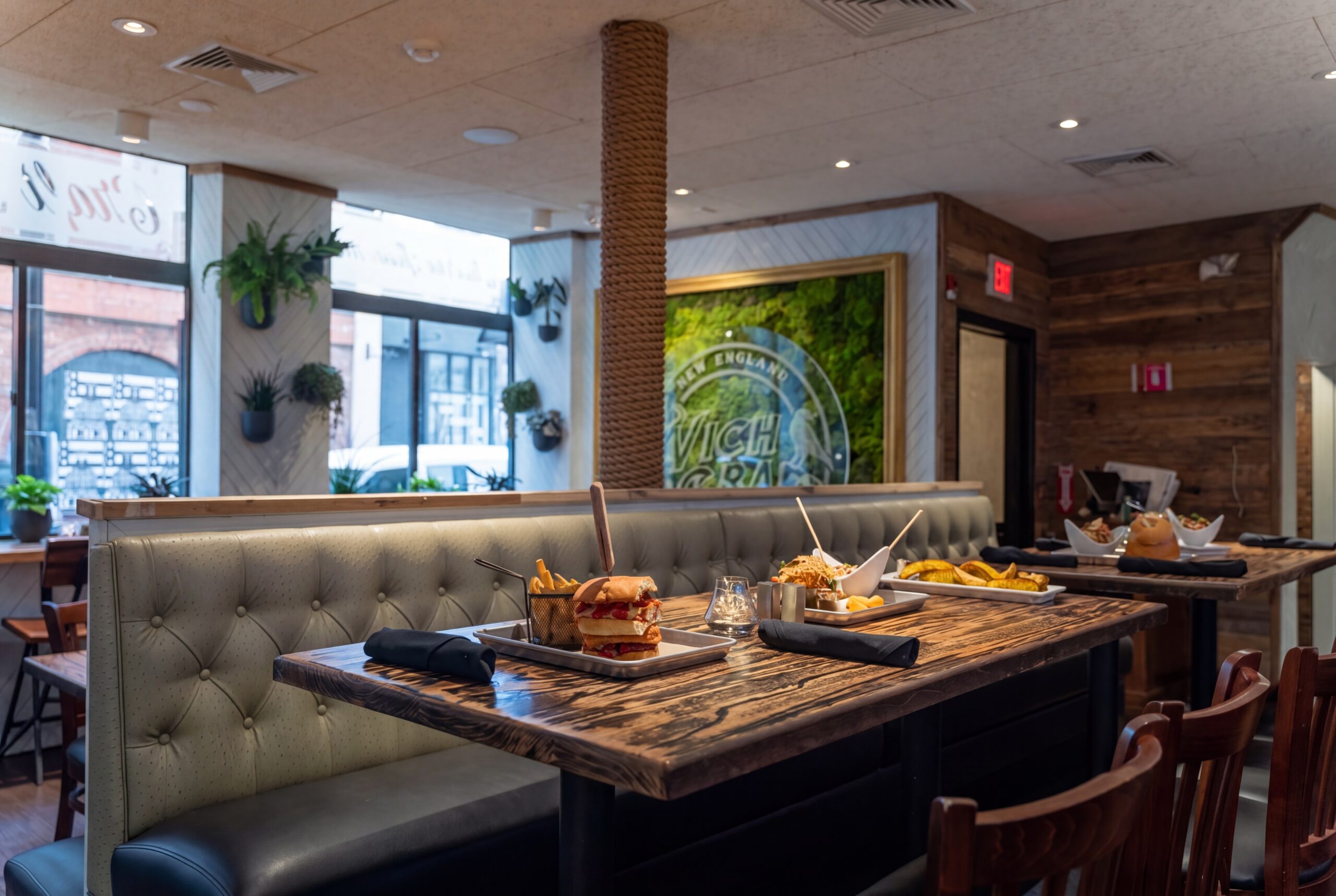 Upscale casual dining setting with tufted banquette seating, rustic wood tables, and plated dishes, complemented by warm lighting, greenery, and a signature moss feature wall<br />
