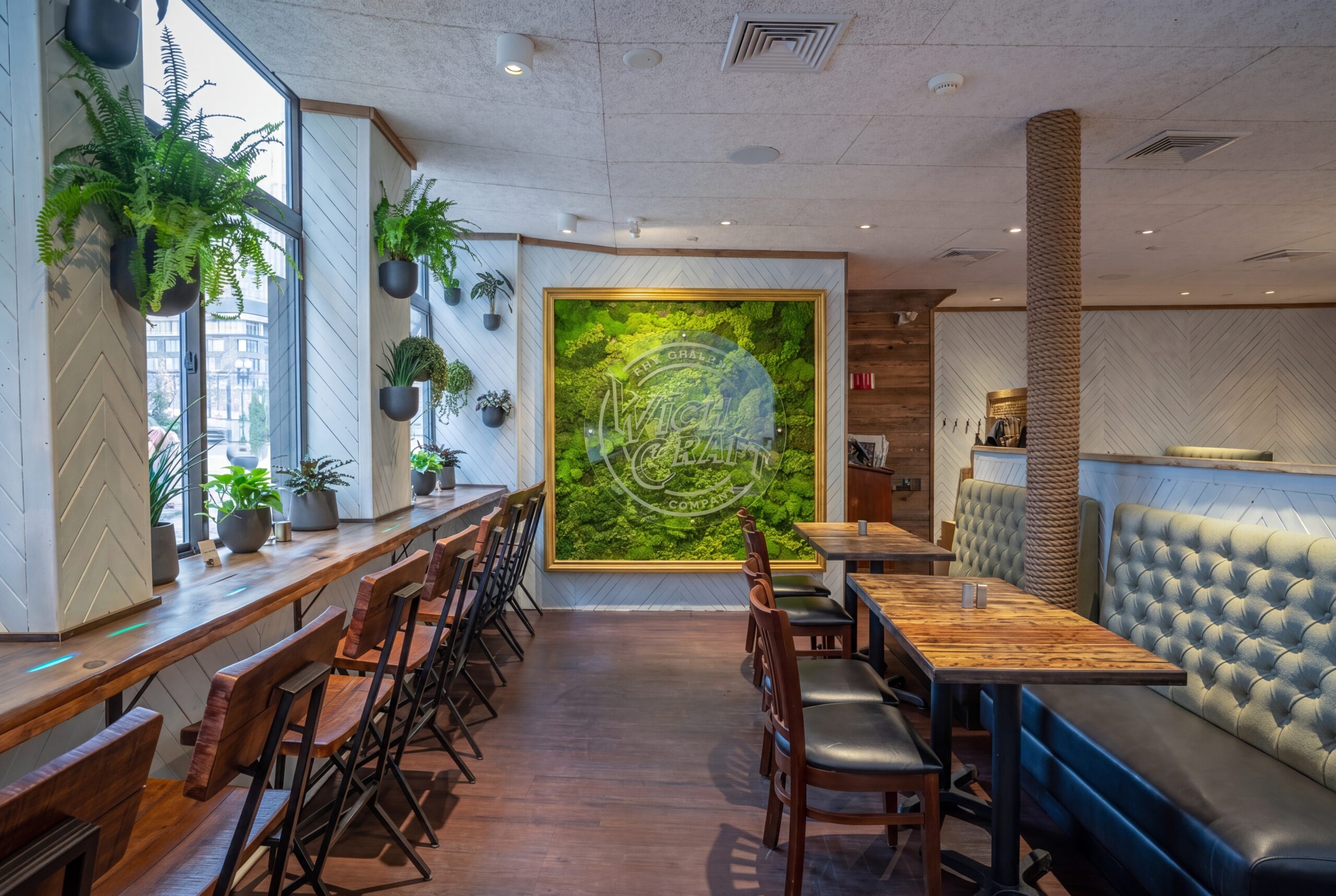 Light-filled restaurant interior with window bar seating, wood stools, lush hanging plants, and a bold moss wall centerpiece creating a fresh, inviting dining atmosphere<br />
