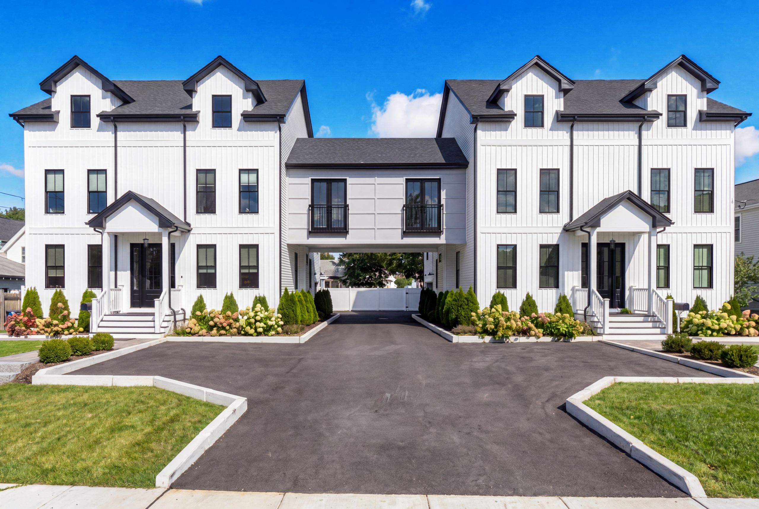 Symmetrical modern farmhouse duplex with white vertical siding, black windows, central breezeway connection, and landscaped front entries along shared driveway<br />

