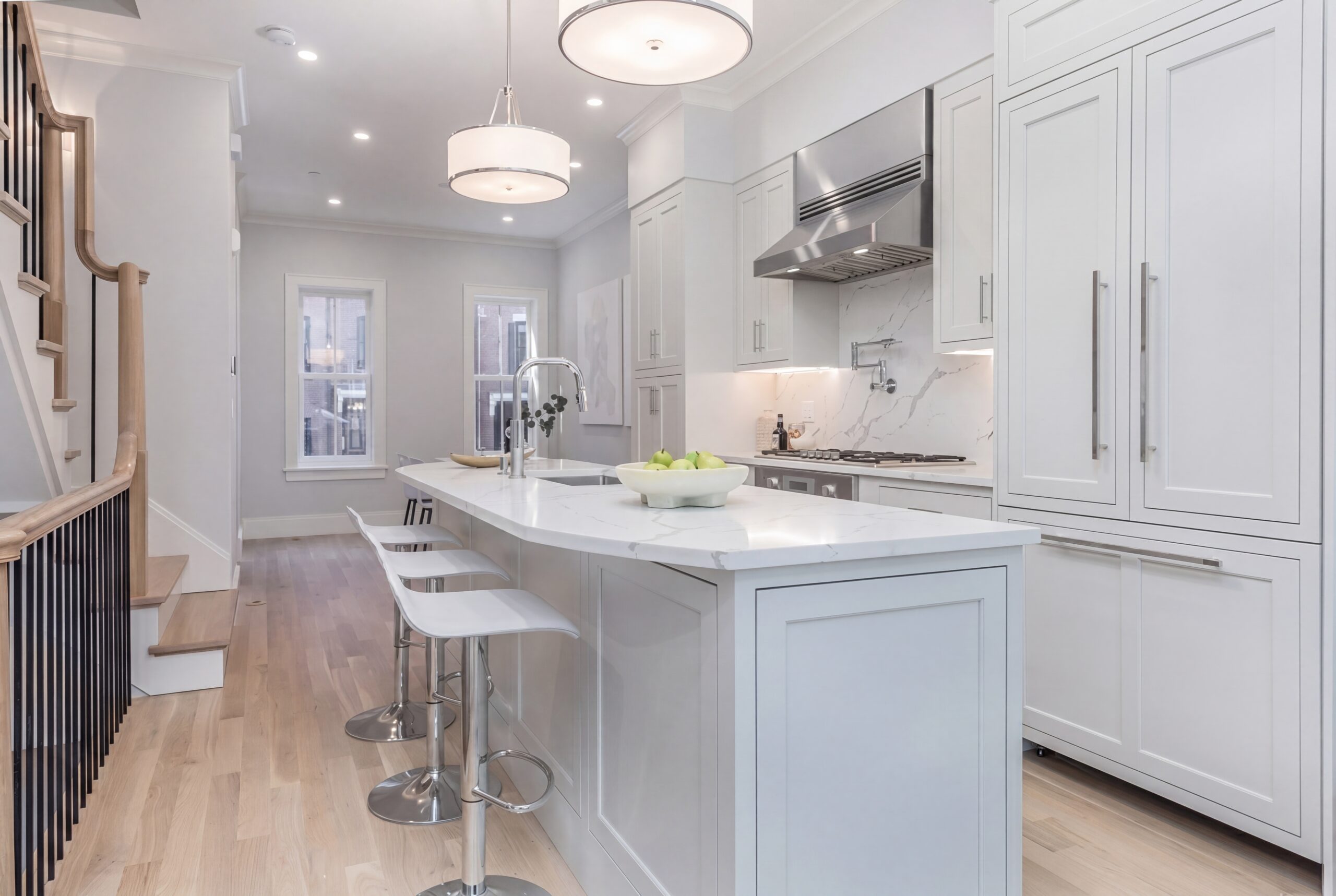 Bright contemporary kitchen interior within a renovated Boston row house, featuring white shaker cabinetry, marble countertops and backsplash, integrated stainless steel appliances, light oak flooring, and a large waterfall island with seating. Soft recessed lighting and modern pendant fixtures create a clean, refined residential atmosphere with an open-concept layout and warm minimalist detailing.<br />

