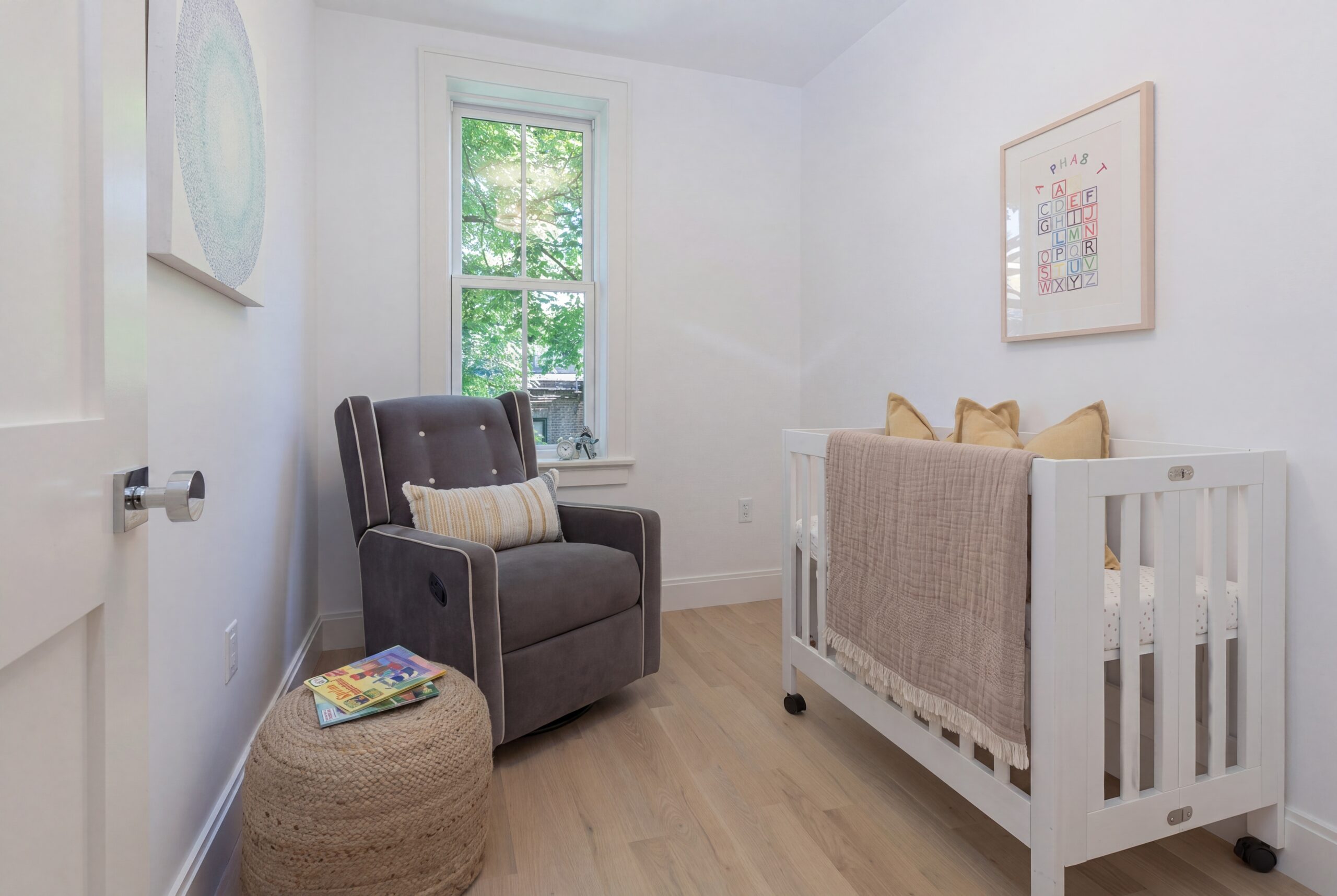 Minimal nursery interior within a renovated Boston row house, featuring a soft neutral palette, light oak flooring, white crib, upholstered rocking chair, and contemporary residential detailing. Natural daylight fills the compact space through a traditional window, creating a calm and inviting atmosphere designed for modern urban family living.<br />
