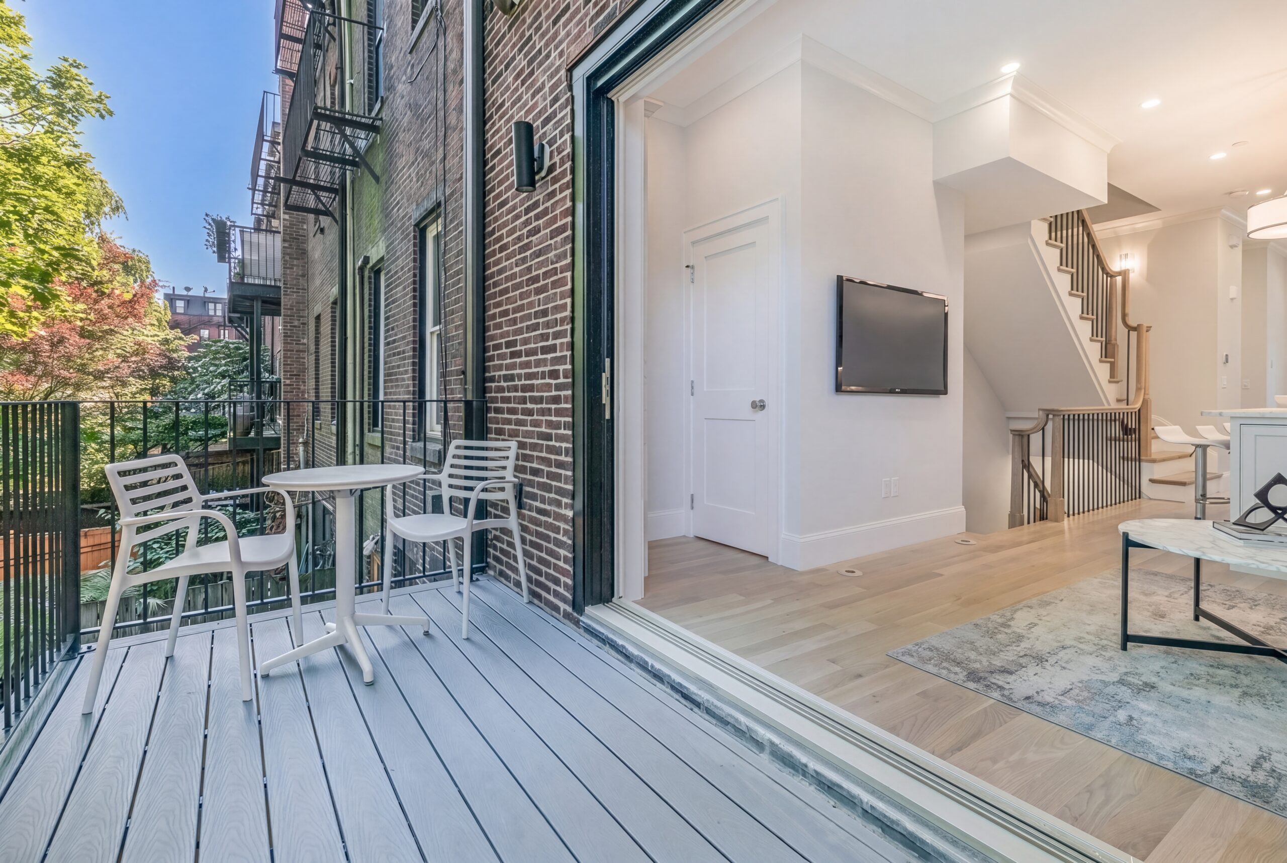 Private outdoor terrace connected to a renovated Boston row house, featuring contemporary decking, minimalist outdoor furnishings, and large sliding glass doors that create a seamless transition between indoor and outdoor living. The adjacent interior showcases bright modern finishes, light oak flooring, and an open residential layout designed for refined urban living.<br />
