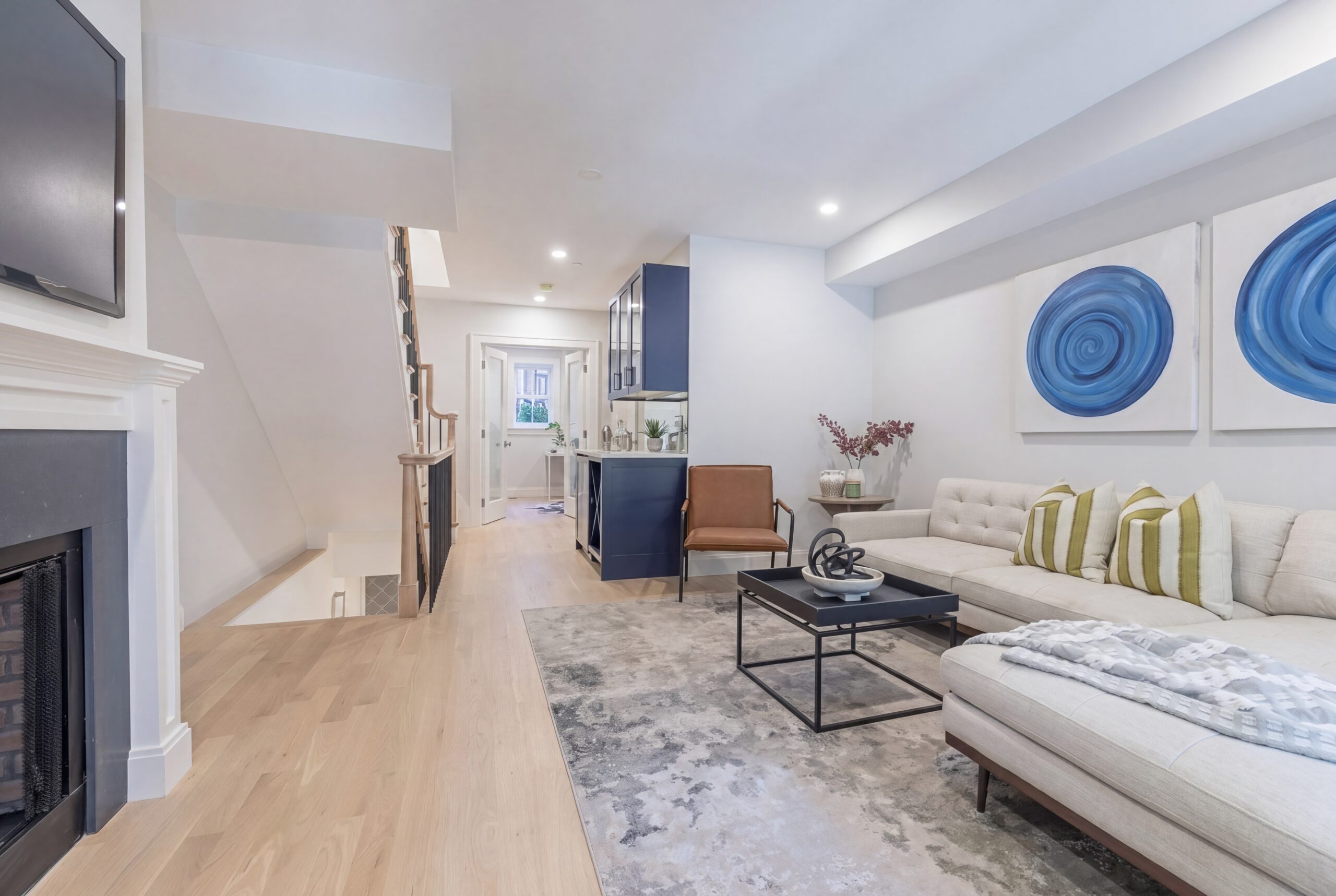 Contemporary lower-level living space within a renovated Boston row house, featuring an open-concept layout with light oak flooring, modern furnishings, recessed lighting, and minimalist interior detailing. A restored fireplace adds historic character while contemporary artwork, clean-lined finishes, and integrated millwork create a refined and comfortable urban residential atmosphere.<br />

