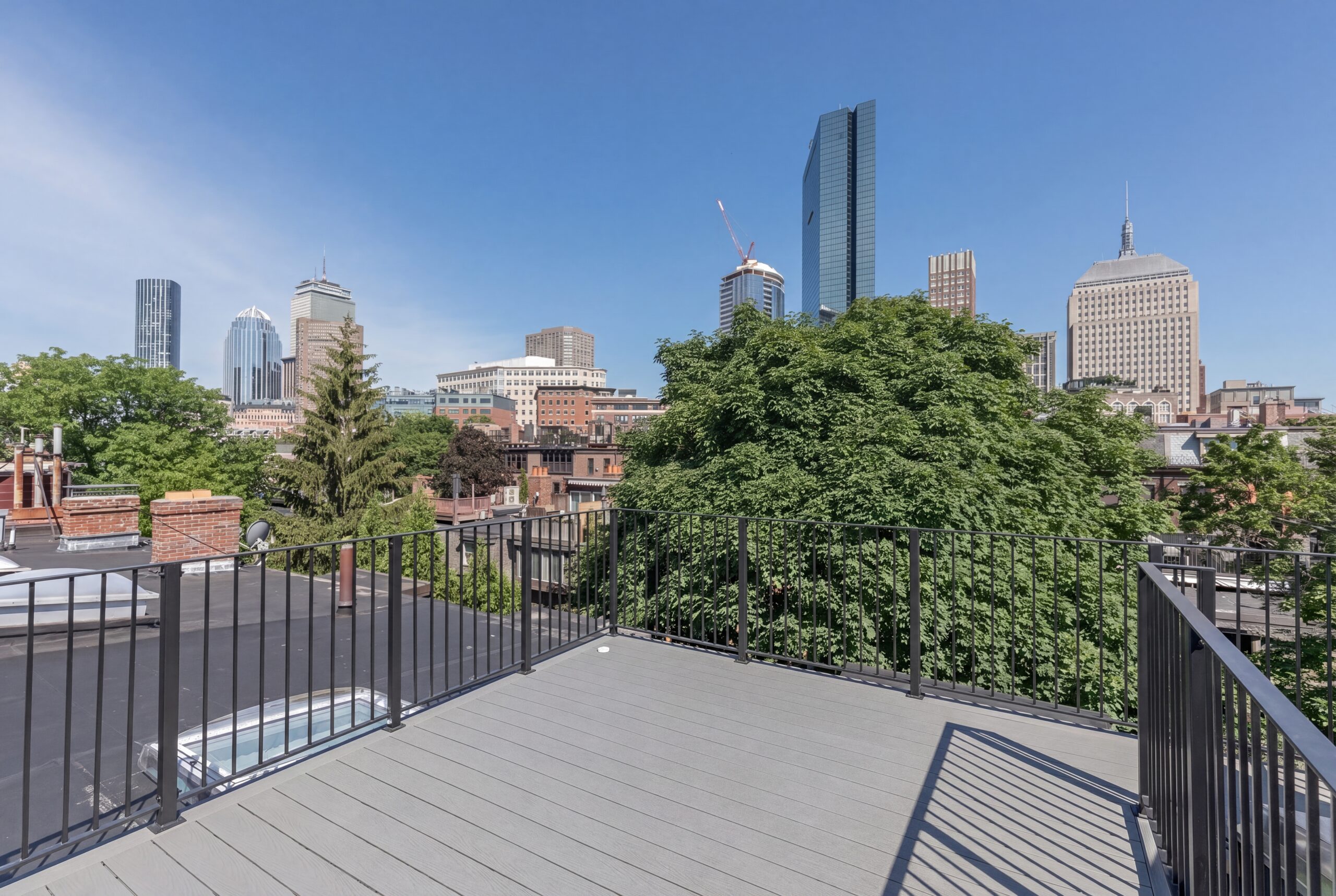 Private rooftop terrace overlooking the Boston skyline, featuring contemporary composite decking, minimalist black metal railings, and panoramic city views framed by mature urban greenery. The outdoor space extends the renovated row house with a refined residential amenity that blends modern outdoor living with the character of Boston’s historic neighborhood context.<br />
