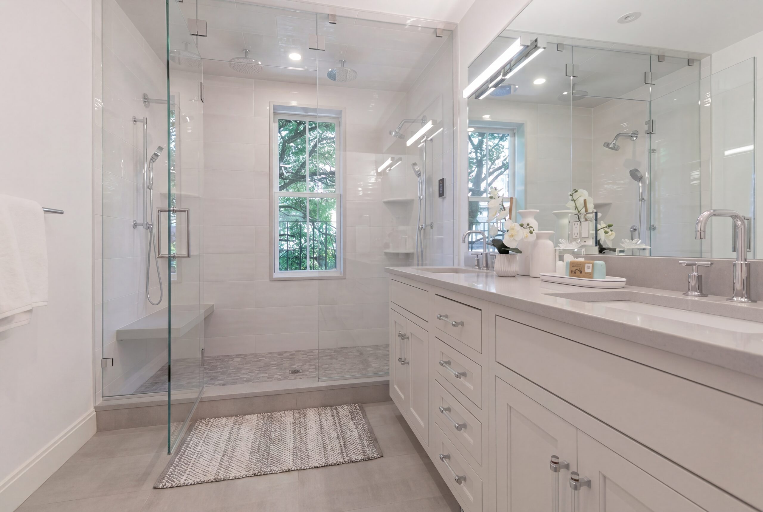 Contemporary primary bathroom within a renovated Boston row house, featuring a bright neutral material palette, custom white vanity cabinetry, large-format tile finishes, and a frameless glass walk-in shower with integrated bench seating. Natural daylight and clean modern detailing create a refined spa-like atmosphere that balances comfort, functionality, and contemporary urban living.<br />
