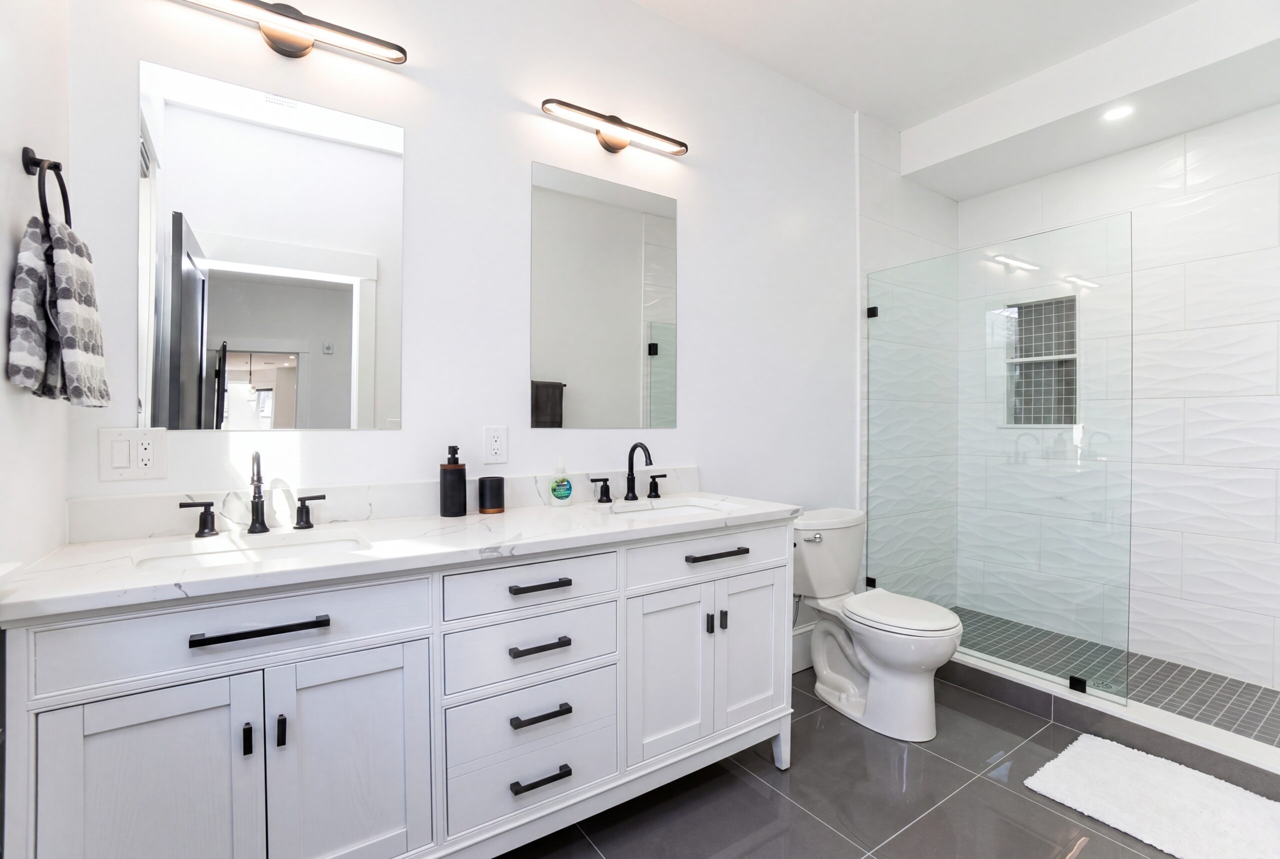 Modern South Boston bathroom renovation with double vanity, black fixtures, frameless glass shower, and contemporary white tile detailing.<br />
