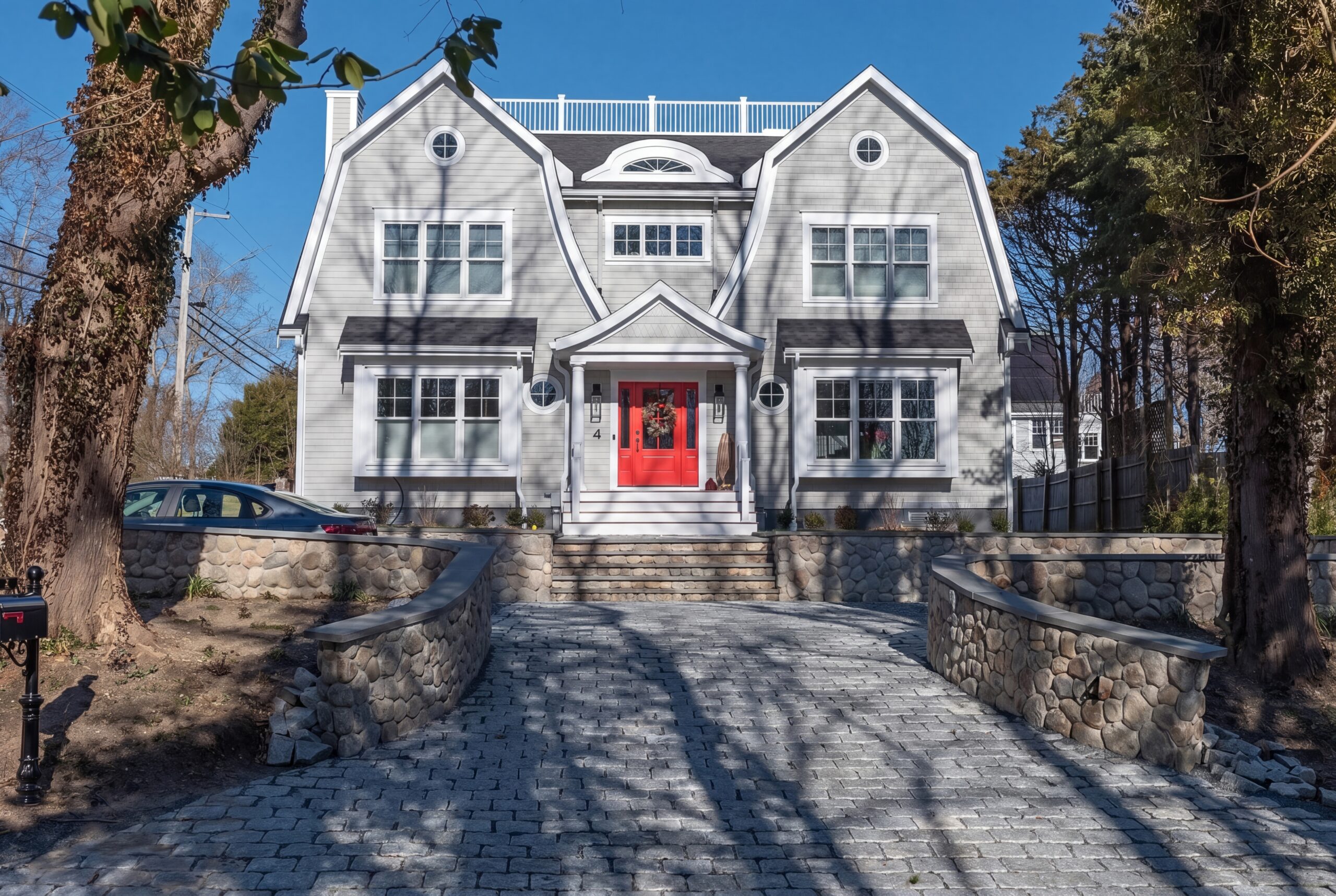 Coastal-inspired single-family home with gambrel roofs, rooftop deck, stone retaining walls, and a bold red front entry door<br />
