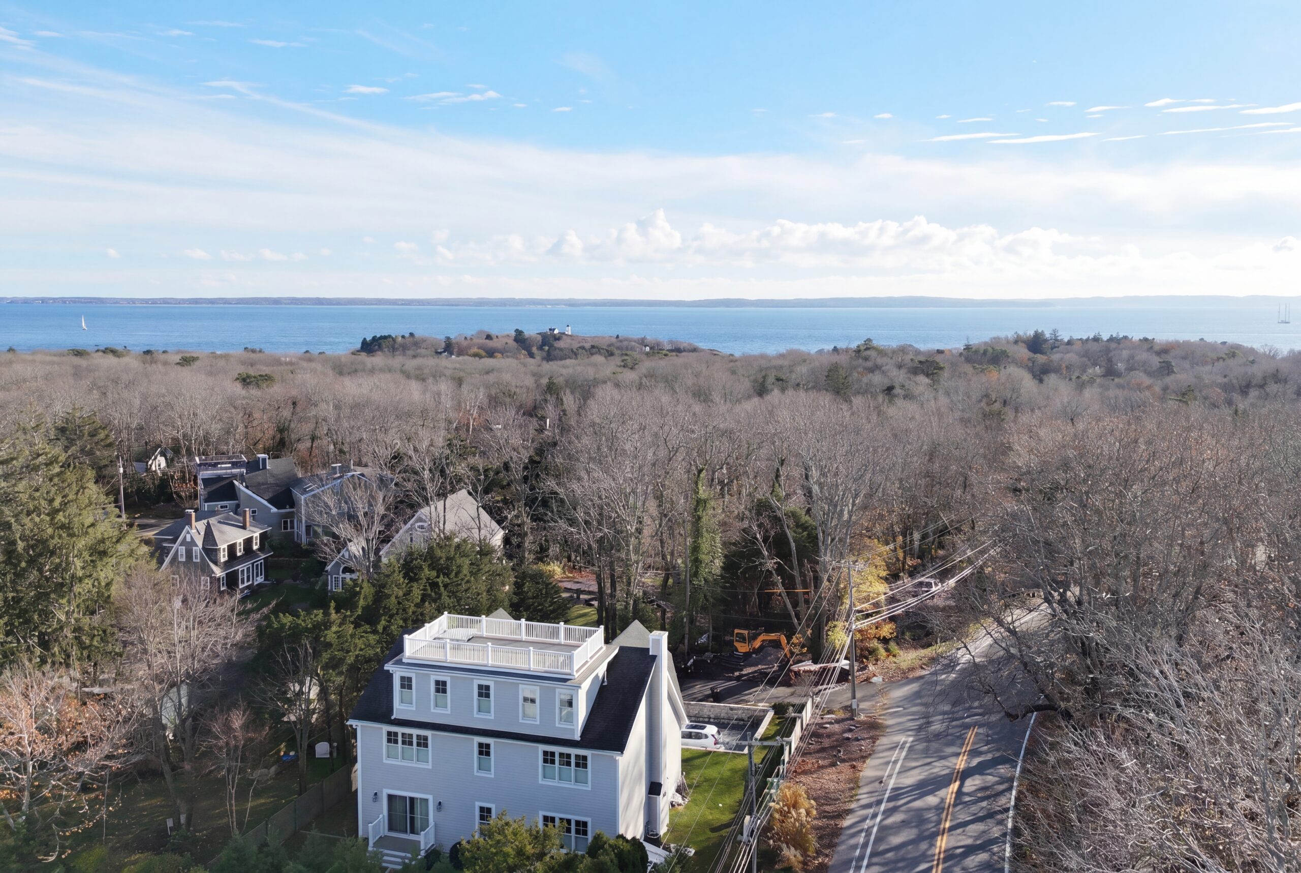 Aerial view of coastal single-family home with rooftop deck overlooking the ocean and surrounding wooded neighborhood landscape<br />
