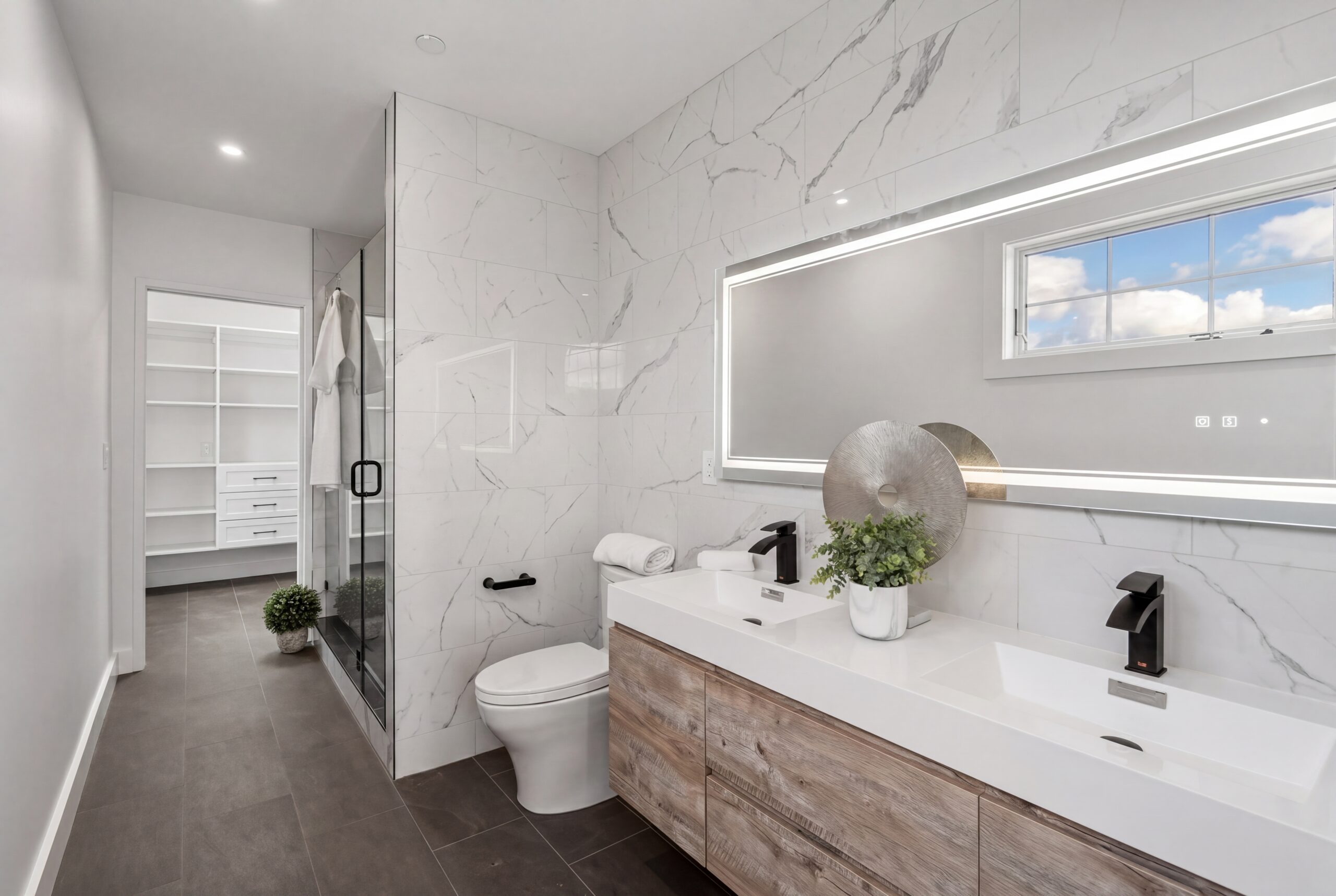Athens St Bathroom Contemporary primary bathroom within a modern multifamily residence, featuring large-format marble-look tile walls, floating wood vanity cabinetry, integrated LED mirror lighting, and matte black plumbing fixtures. A frameless glass shower enclosure and adjacent walk-in closet create a refined spa-like atmosphere that combines minimalist modern detailing with luxury urban residential living.<br />