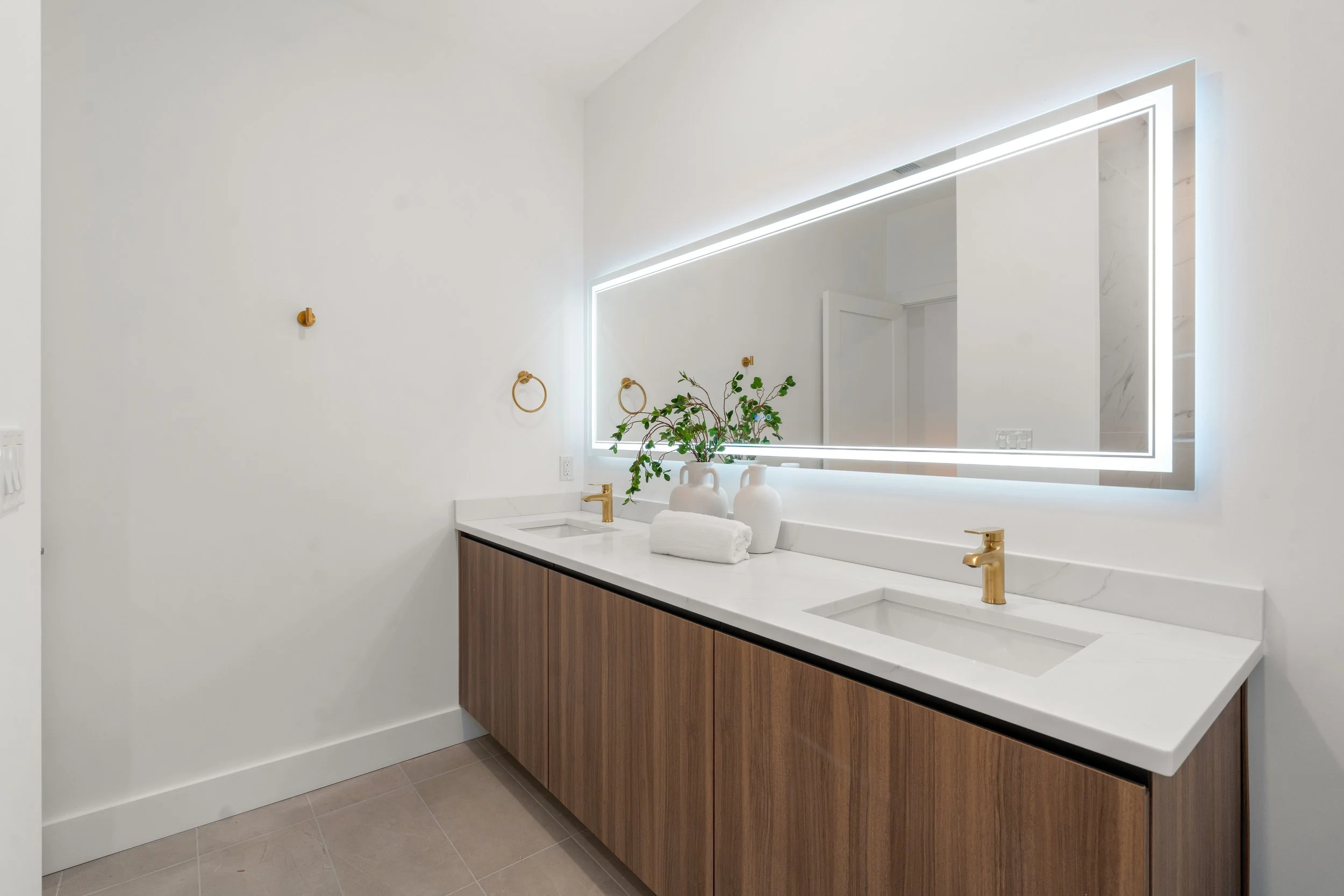 Contemporary bathroom with double vanity, wood cabinetry, brass fixtures, and large backlit mirror
