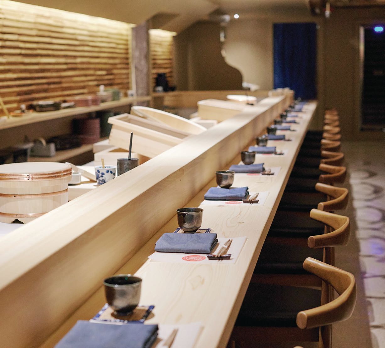 Minimalist sushi counter featuring a custom light wood bar, warm ambient lighting, and intimate seating arranged for an immersive omakase dining experience.<br />
