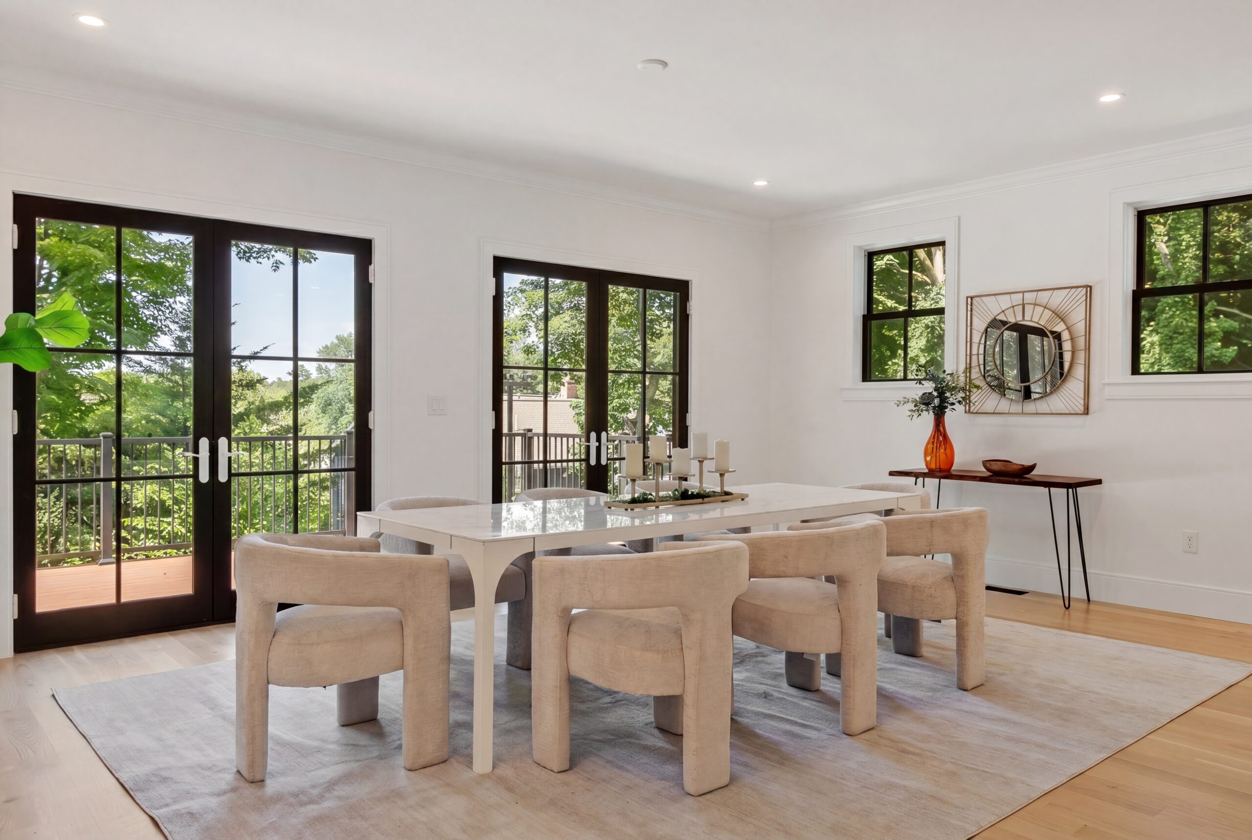 Bright contemporary dining room featuring black-framed French doors, oversized windows, light wood flooring, modern upholstered seating, and seamless indoor-outdoor living with abundant natural light.
