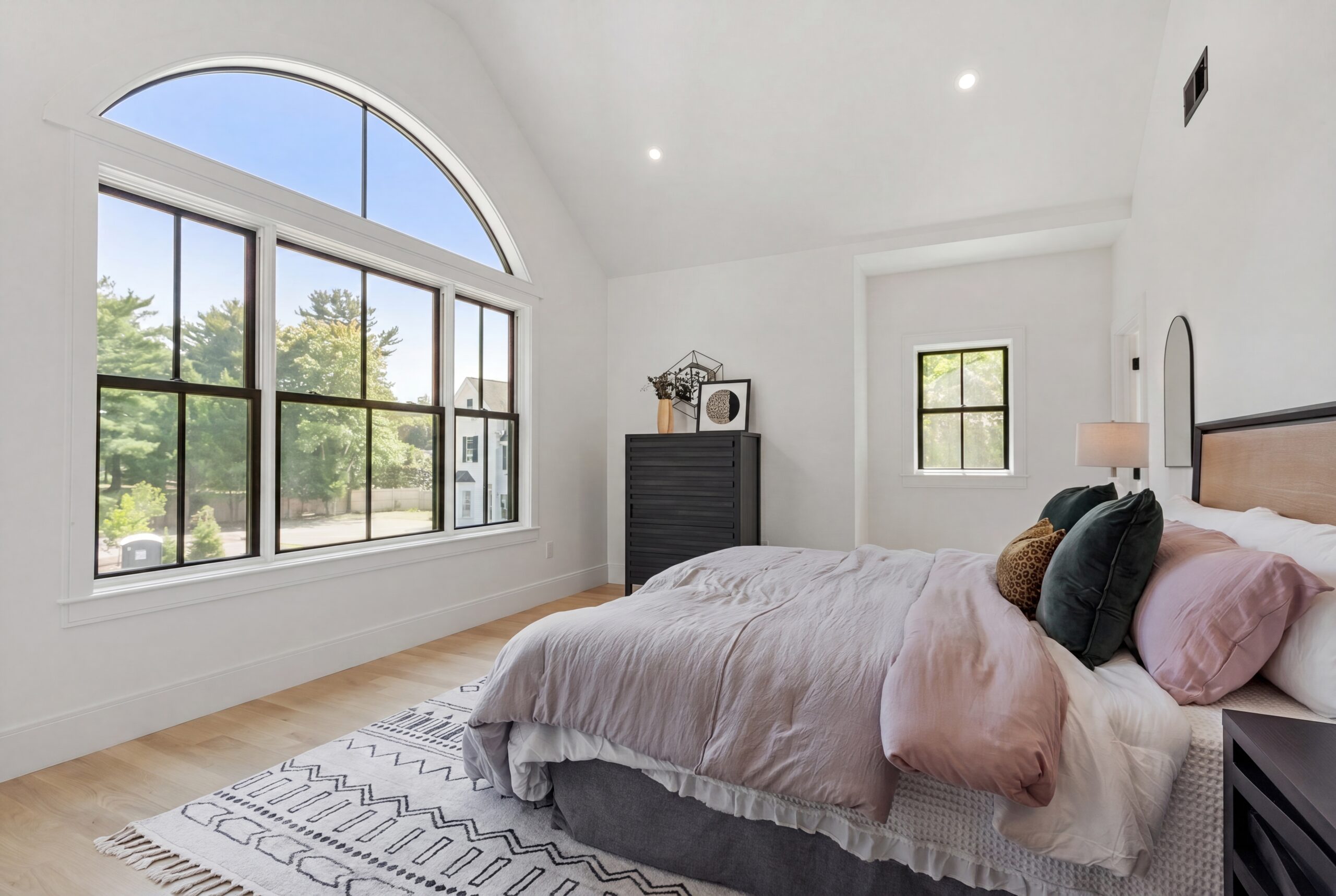 Primary bedroom featuring a vaulted ceiling, oversized arched window, abundant natural light, light wood flooring, and contemporary finishes in a luxury single-family residence.