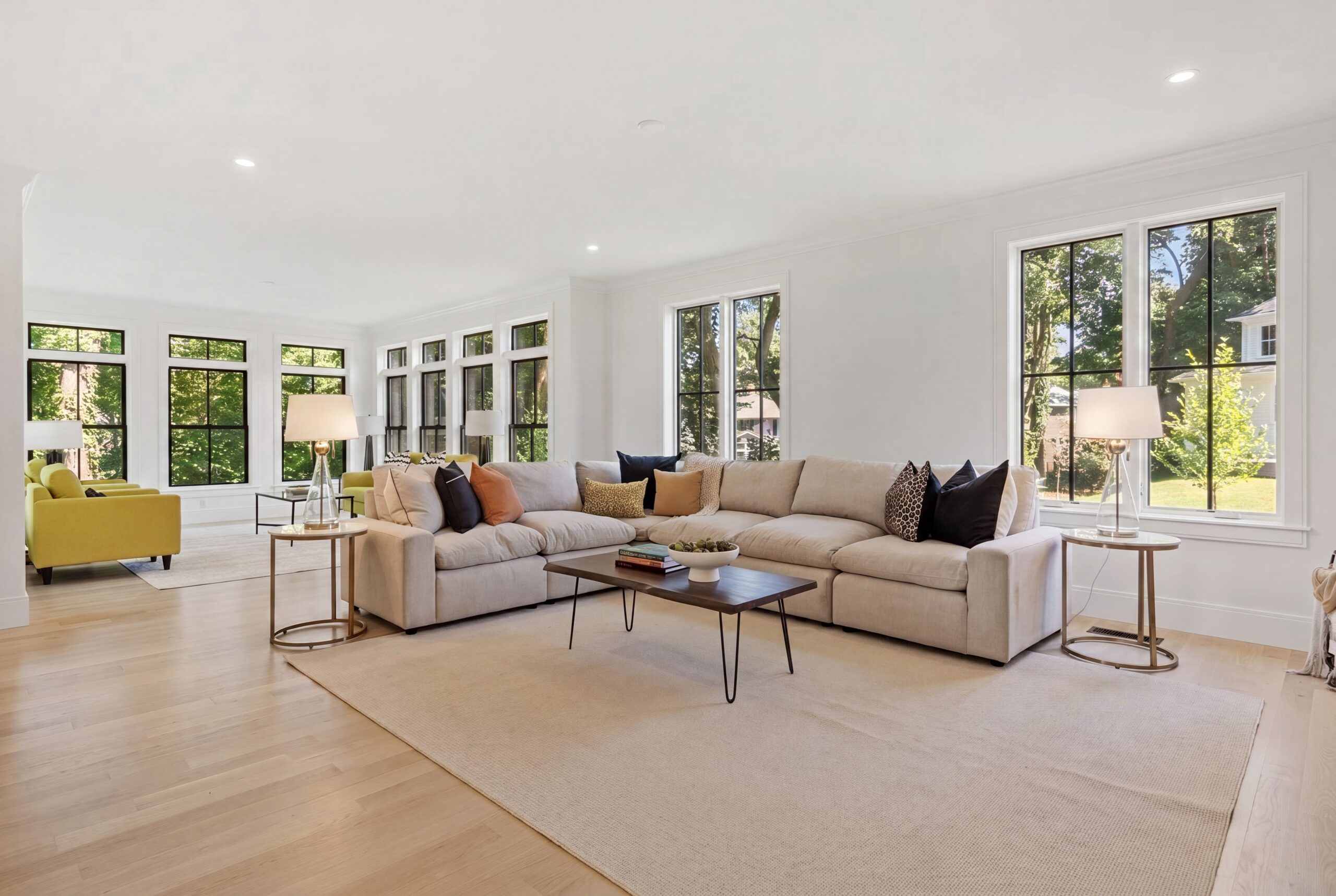 Open-concept living room featuring oversized black-framed windows, abundant natural light, light wood flooring, contemporary furnishings, and seamless connection to a sunroom in a luxury single-family residence.