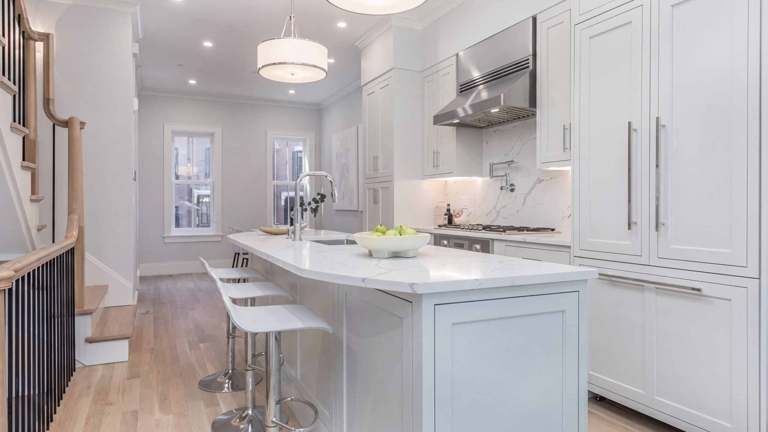 Bright contemporary kitchen interior within a renovated Boston row house, featuring white shaker cabinetry, marble countertops and backsplash, integrated stainless steel appliances, light oak flooring, and a large waterfall island with seating. Soft recessed lighting and modern pendant fixtures create a clean, refined residential atmosphere with an open-concept layout and warm minimalist detailing.