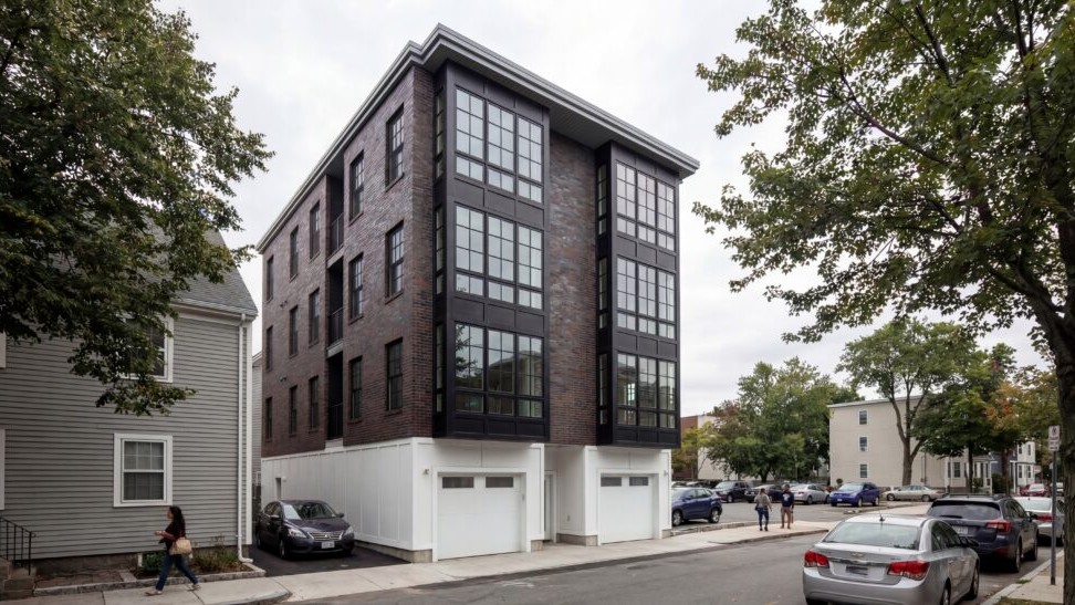 athens st front 16-9 Contemporary multifamily residential building featuring a dark brick façade, expansive black-framed window walls, and modern urban detailing within a dense neighborhood streetscape. The four-story development combines clean contemporary forms with warm masonry materials, integrated garage access, and large vertical glazing elements that create a bold architectural presence against the surrounding residential context.