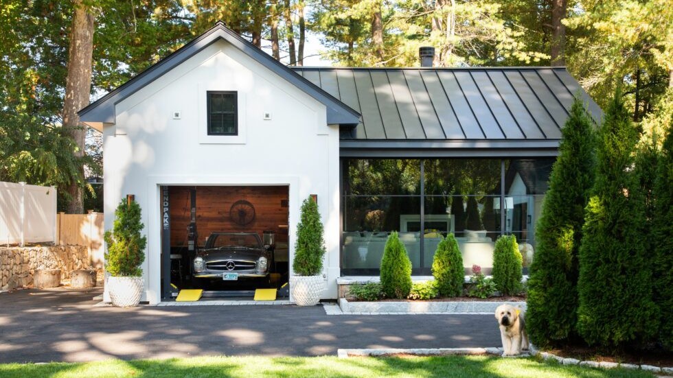 Modern garage addition with white exterior and black metal roof, featuring a glass-walled living space and a vintage car showcased at the entry, set within a landscaped residential setting.